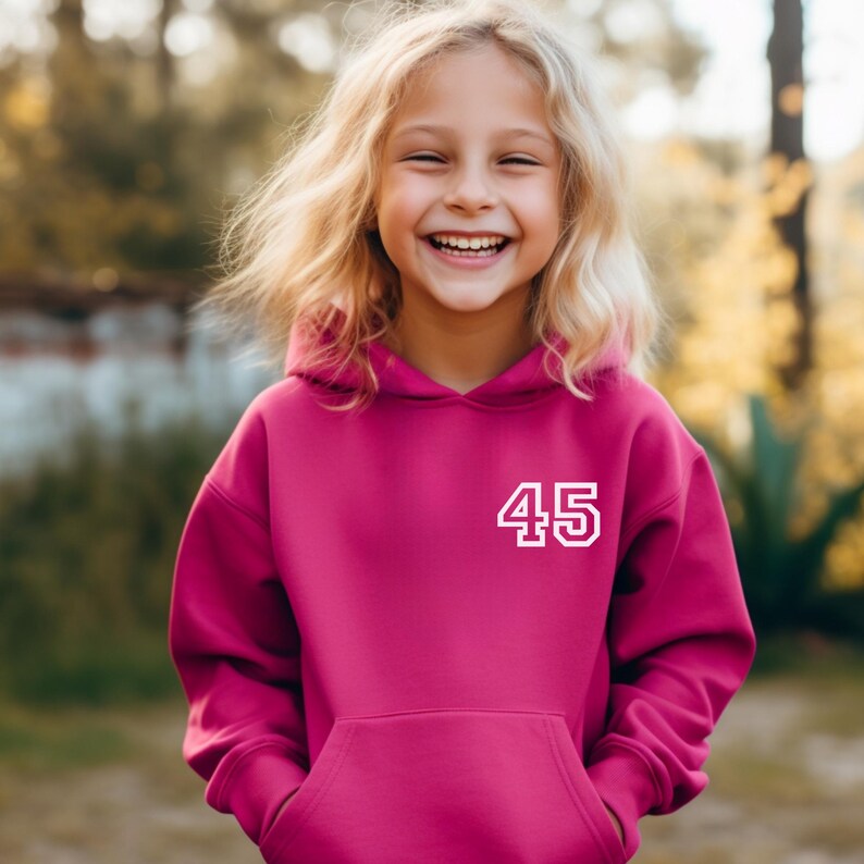 May include: A magenta hoodie with a white number "45" on the front. The hoodie has a kangaroo pocket and a hood. The person wearing the hoodie is smiling with blonde hair.