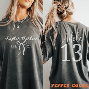 May include: A charcoal grey short-sleeve t-shirt featuring white text and graphics. The front displays the words chapter thirteen, EST. 2013, and a bow icon. The back features the name Adele above the large number 13 in white lettering.