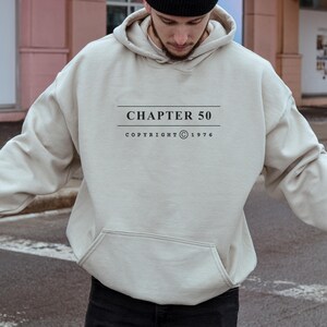 May include: A beige hooded sweatshirt featuring a front kangaroo pocket and black text printed on the chest that reads CHAPTER 50 COPYRIGHT 1976, framed by two horizontal lines above and below the main text.