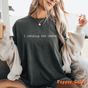 May include: Dark gray t-shirt with the text "I catalog the chaos" in white. The shirt is a relaxed fit and has a vintage wash. The words "Pepper Color" are in orange at the bottom right.