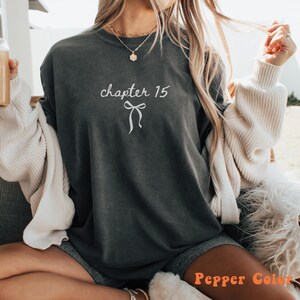 May include: Dark gray t-shirt with the text "chapter 15" and a bow graphic in white. The shirt is a pepper color and has a relaxed fit. The shirt is a casual wear item.
