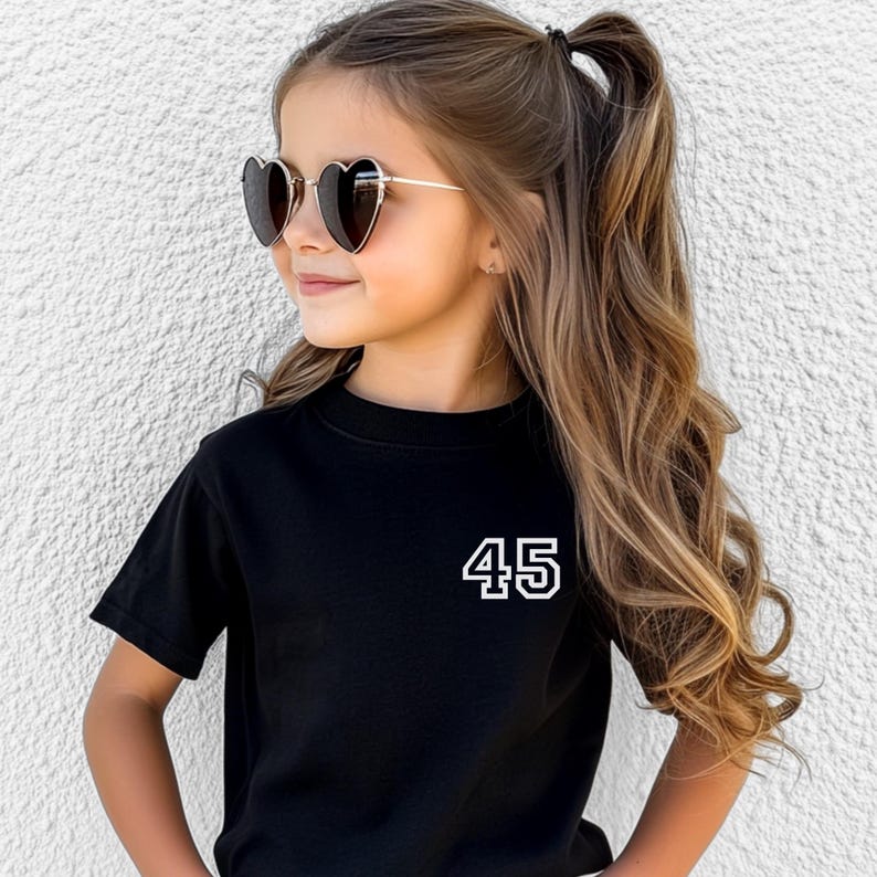 May include: A black t-shirt with the number "45" in white. The shirt is worn with heart-shaped sunglasses with gold frames. The child has long, wavy brown hair.