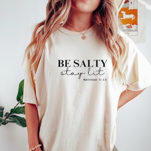 May include: Cream-colored t-shirt with the text "BE SALTY stay lit" in black script. The Bible verse Matthew 5:13 is printed below the phrase. The shirt is a casual, oversized fit.