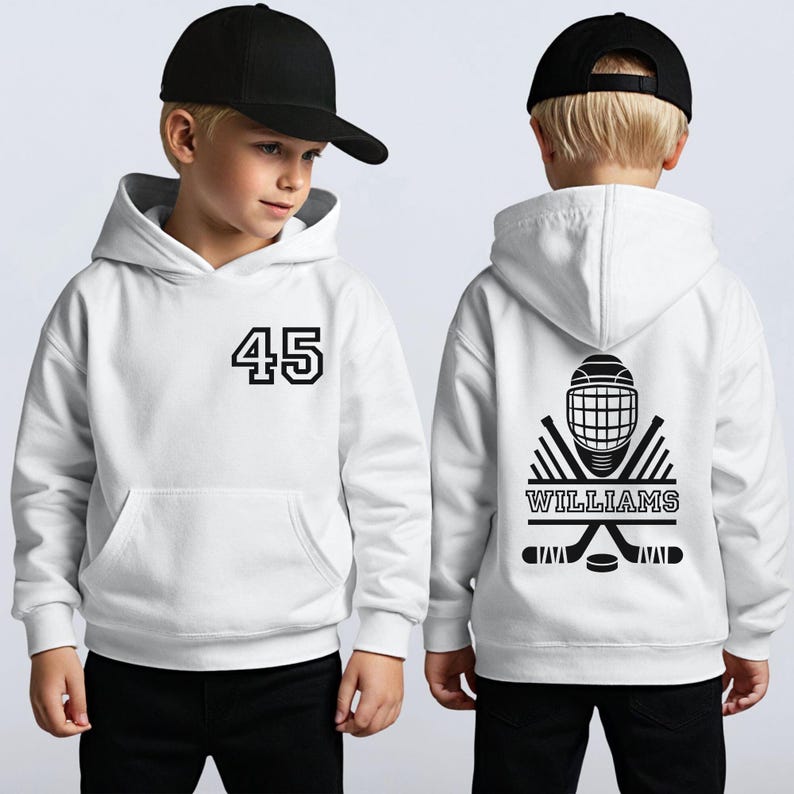 May include: White hoodie with a front pocket, featuring the number 45 in black. The back displays a hockey-themed graphic with a helmet, crossed sticks, a puck, and the name "WILLIAMS" in a banner. The hoodie is worn by a child.