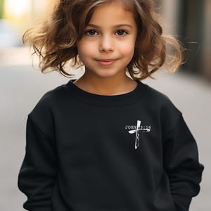 K&ouml;nnte beinhalten: Schwarzer Pullover mit einem wei&szlig;en Kreuz und dem Text "JOHN 3:16" auf der Vorderseite.
