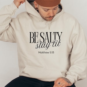 May include: A beige hoodie with the text "BE SALTY stay lit" in black script and block letters, with "Matthew 5:13" below. The hoodie has a drawstring and a front pocket.