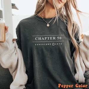 May include: A charcoal gray crewneck sweatshirt featuring the white text "CHAPTER 50" centered between two horizontal lines, with the text "COPYRIGHT © 1976" printed directly below. The garment has a vintage-washed appearance and long sleeves.