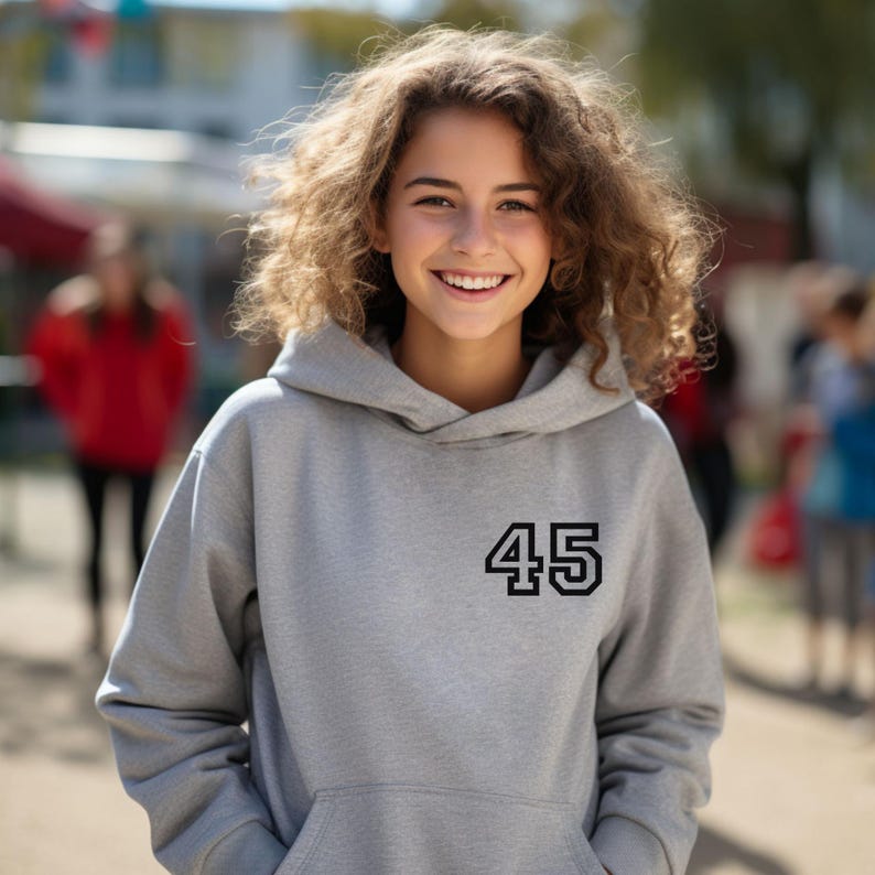 May include: A gray hooded sweatshirt with the number 45 in black on the front. The sweatshirt has a drawstring hood and a front pocket. The person wearing the sweatshirt is smiling.