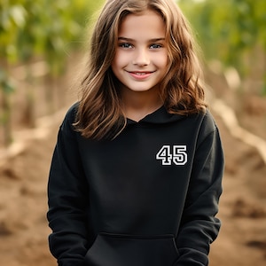 May include: A black hooded sweatshirt with the number "45" in white block letters. The sweatshirt has a front pocket and a drawstring hood. The background is blurred, with a young person in the foreground.