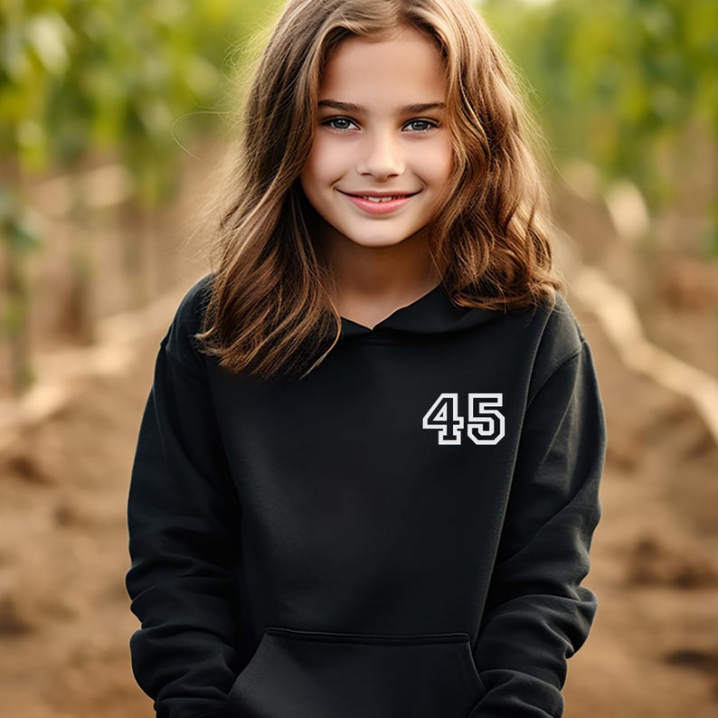 May include: A black hooded sweatshirt with the number "45" in white block letters. The sweatshirt has a front pocket and a drawstring hood. The background is blurred, with a young person in the foreground.