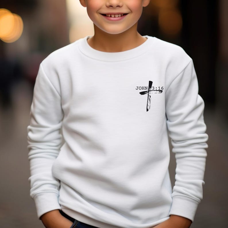 K&ouml;nnte beinhalten: Wei&szlig;er Langarm-Pullover mit einem schwarzen Kreuz und dem Text "JOHN 3:16" auf der Vorderseite.