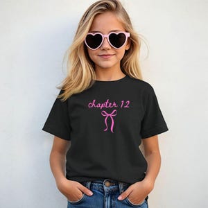 May include: Black t-shirt with the text "chapter 12" in pink script, accompanied by a pink bow graphic. The shirt is worn by a person wearing pink heart-shaped sunglasses.
