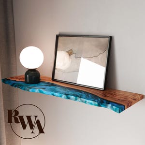 May include: A floating wooden shelf with a live edge and a blue resin inlay. A black framed picture and a small lamp with a white globe are on the shelf. The shelf is mounted on a white wall.