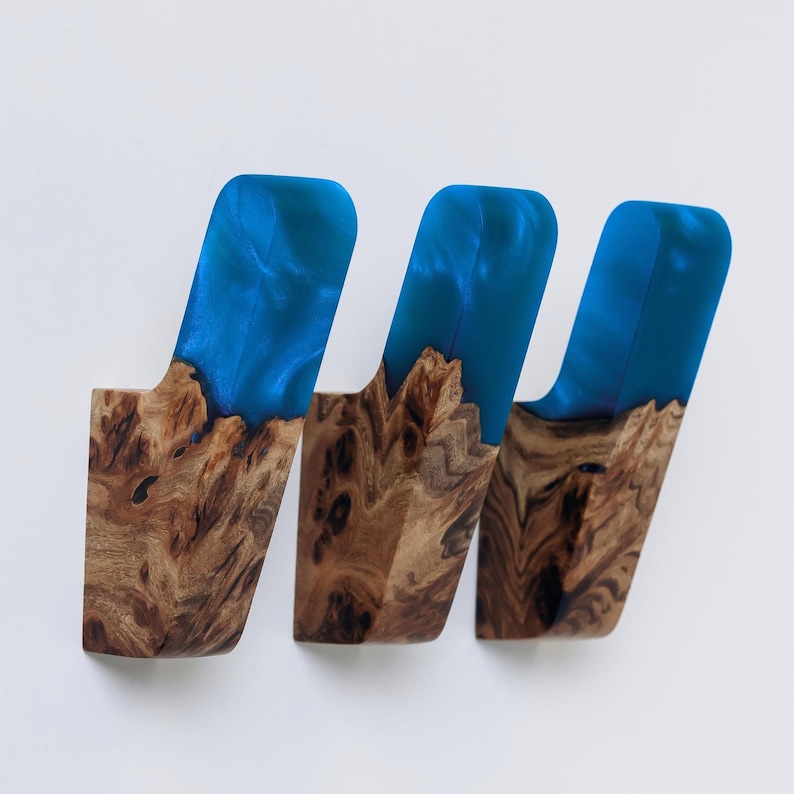 Op de afbeelding: Drie wandhaken met een uniek ontwerp. Elke haak heeft een houten basis met een natuurlijke nerf, bekroond met levendig blauwe hars. De hars heeft een glanzende afwerking, wat een opvallend contrast met het hout cre&euml;ert.