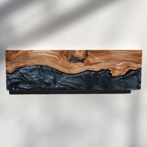 Puede incluir: Un estante de pared rectangular que presenta una secci&oacute;n superior de madera natural de color marr&oacute;n claro con un nudo prominente y una secci&oacute;n inferior contrastante de resina negra lisa con una textura veteada y arremolinada.