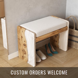 Può includere: Una panca bianca e in legno con un sedile imbottito. La panca ha un design unico con un inserto in resina bianca. La panca è seduta su un pavimento in legno con le scarpe sotto.