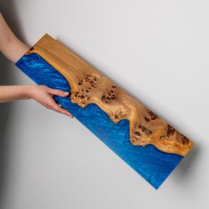 Puede incluir: Una tabla de servir rectangular que presenta una secci&oacute;n de madera natural con borde vivo, nudos oscuros y vetas marcadas, unida a una secci&oacute;n de resina epoxi azul transl&uacute;cida y brillante con un dise&ntilde;o de remolinos.