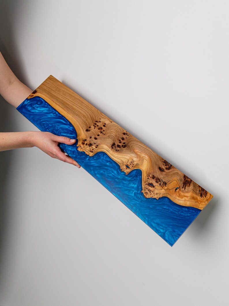 Puede incluir: Una tabla de servir rectangular que presenta una secci&oacute;n de madera natural con borde vivo, nudos oscuros y vetas marcadas, unida a una secci&oacute;n de resina epoxi azul transl&uacute;cida y brillante con un dise&ntilde;o de remolinos.