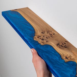 Puede incluir: Un estante rectangular hecho de una losa de madera natural con un patr&oacute;n de grano nudoso prominente en un lado y una secci&oacute;n de resina epoxi azul transl&uacute;cida y arremolinada en el otro, creando una superficie lisa y continua con un dise&ntilde;o fluvial.
