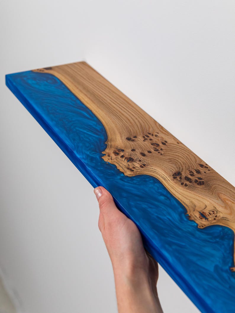 Puede incluir: Un estante rectangular hecho de una losa de madera natural con un patr&oacute;n de grano nudoso prominente en un lado y una secci&oacute;n de resina epoxi azul transl&uacute;cida y arremolinada en el otro, creando una superficie lisa y continua con un dise&ntilde;o fluvial.