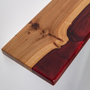 Puede incluir: Una tabla de servir rectangular que presenta una secci&oacute;n de madera de color marr&oacute;n claro natural con nudos visibles en un lado y una secci&oacute;n de resina roja transl&uacute;cida profunda y lisa en el otro, unidas por un patr&oacute;n org&aacute;nico ondulado a lo largo.