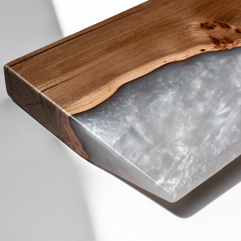 Puede incluir: Una tabla de servir rectangular que presenta un borde de madera marr&oacute;n natural con veta visible y una secci&oacute;n de resina blanca semitransl&uacute;cida con una textura marmolada nacarada que contrasta con la l&iacute;nea org&aacute;nica y ondulada de la madera.