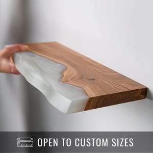 May include: A rectangular wooden shelf with a live edge design, featuring a light-colored epoxy resin. The shelf is a natural wood tone with a smooth, polished finish. The text "OPEN TO CUSTOM SIZES" is displayed.