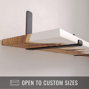 Puede incluir: Estantería flotante con parte superior blanca e inferior de vetas de madera, soportada por soportes metálicos negros. La estantería está montada en una pared blanca. El texto de la imagen dice "OPEN TO CUSTOM SIZES".