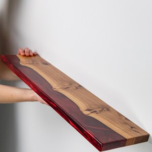 Puede incluir: Un estante flotante rectangular que presenta una secci&oacute;n de madera de color marr&oacute;n claro natural con nudos visibles y una secci&oacute;n de resina transl&uacute;cida de color rojo intenso que recorre un lado, creando una superficie plana y lisa.