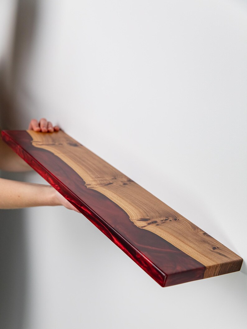Puede incluir: Un estante flotante rectangular que presenta una secci&oacute;n de madera de color marr&oacute;n claro natural con nudos visibles y una secci&oacute;n de resina transl&uacute;cida de color rojo intenso que recorre un lado, creando una superficie plana y lisa.