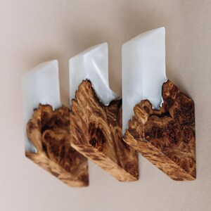 Op de afbeelding: Drie houten wandkunstwerken. Elk stuk heeft een lichtgekleurd, rechthoekig bovendeel en een getextureerde, bruine houten basis. De houtnerf is zichtbaar. De stukken zijn op een neutraal gekleurde muur gemonteerd. Uniek ontwerp.
