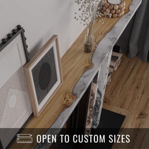 Puede incluir: Una repisa de madera de tamaño personalizado con una incrustación de mármol y resina. La repisa presenta un patrón de vetas de madera natural y está decorada con un jarrón de flores blancas, un candelabro y obras de arte enmarcadas. El texto en la imagen dice "OPEN TO CUSTOM SIZES."