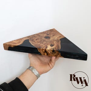 May include: A triangular shelf made of natural wood burl with a prominent grain pattern and smooth, dark black resin filling the gaps. The piece features a sharp geometric shape with a polished finish and includes the letters RWA in a circular logo.