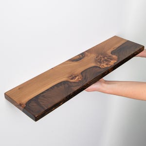 Puede incluir: Un estante flotante rectangular de madera con un dise&ntilde;o de borde natural. La madera muestra un patr&oacute;n de veta contrastante con secciones de color tostado claro y detalles de madera nudosa de color marr&oacute;n oscuro en la superficie.