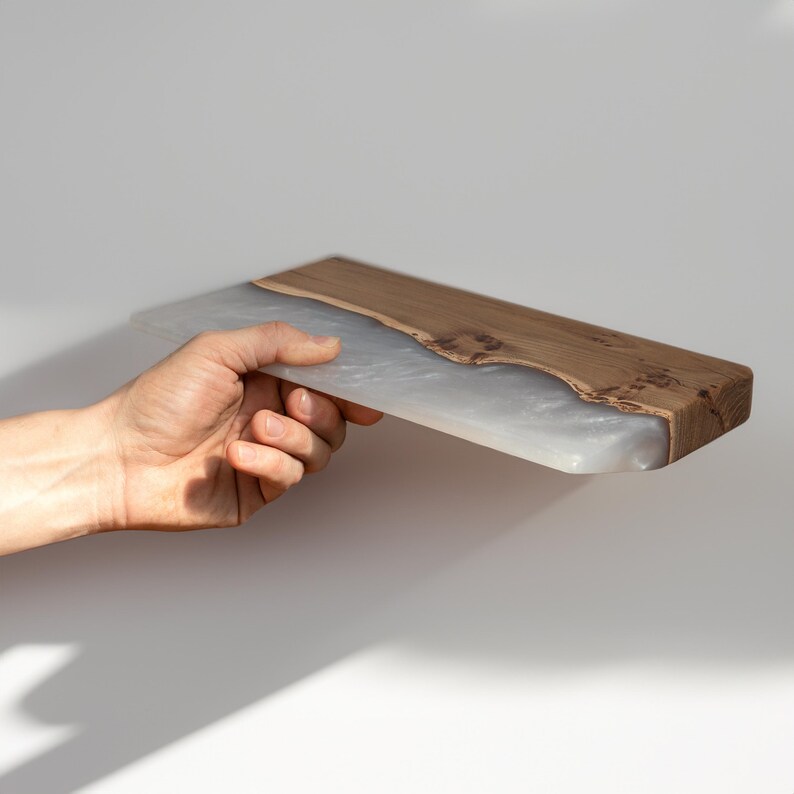 Puede incluir: Una tabla de servir rectangular que presenta una secci&oacute;n de madera natural con un patr&oacute;n de grano visible y una secci&oacute;n de resina blanca transl&uacute;cida en contraste con una textura marmolada y arremolinada. La tabla tiene una superficie lisa.