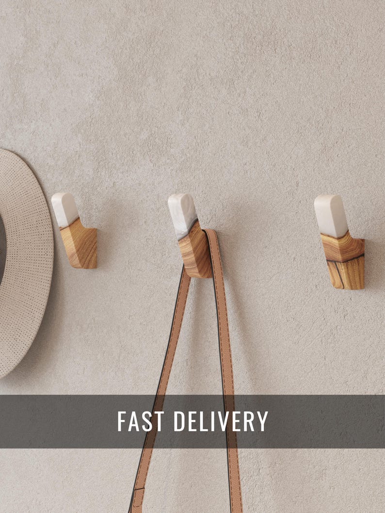 Op de afbeelding: Drie houten wandhaken met een lichte afwerking en witte accenten. Een bruinleren riem hangt aan &eacute;&eacute;n haak. De haken zijn bevestigd aan een lichtgrijze muur. De tekst "FAST DELIVERY" wordt weergegeven.
