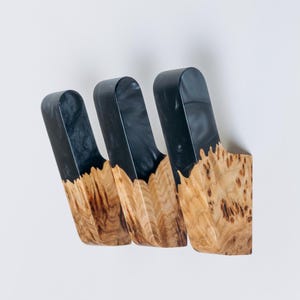 Op de afbeelding: Drie aan de muur gemonteerde houten haken. Het bovenste deel van elke haak is donkergrijs, terwijl het onderste deel een natuurlijke houtnerf heeft. De haken zijn aan een witte muur bevestigd.