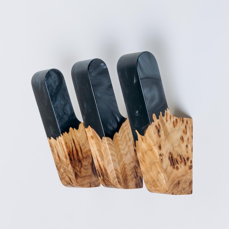 Op de afbeelding: Drie aan de muur gemonteerde houten haken. Het bovenste deel van elke haak is donkergrijs, terwijl het onderste deel een natuurlijke houtnerf heeft. De haken zijn aan een witte muur bevestigd.