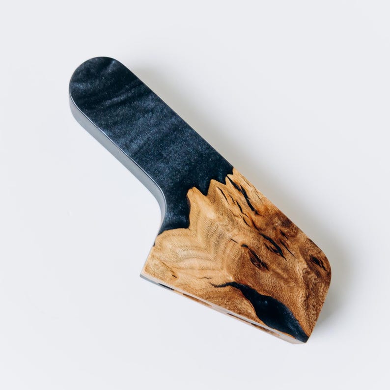 Op de afbeelding: Een handgemaakt kaasmes met een houten handvat en een donkerblauwe harsinleg. Het handvat is gemaakt van natuurlijk hout met een uniek nerfpatroon. Het mes is ontworpen voor het snijden van kaas en andere zachte voedingsmiddelen.