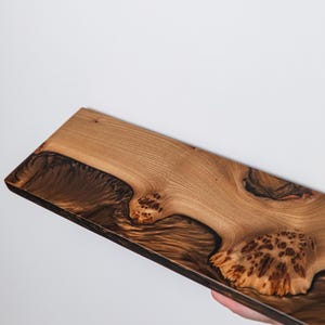 Puede incluir: Una tabla de madera rectangular que presenta una veta de madera natural de color marr&oacute;n claro contrastada con secciones de resina oscura y arremolinada. La superficie muestra nudos de madera org&aacute;nicos y patrones complejos con un acabado pulido.