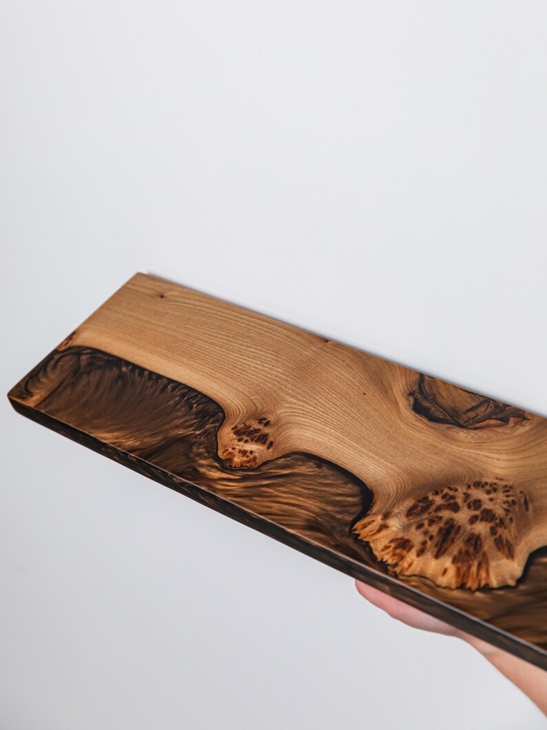 Puede incluir: Una tabla de madera rectangular que presenta una veta de madera natural de color marr&oacute;n claro contrastada con secciones de resina oscura y arremolinada. La superficie muestra nudos de madera org&aacute;nicos y patrones complejos con un acabado pulido.