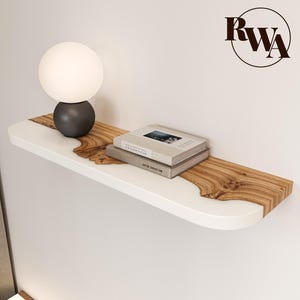 May include: A wall-mounted floating shelf featuring a white matte finish with a central inlay of natural wood grain. A spherical white glass lamp with a dark base and two stacked hardcover books are placed on the shelf surface.