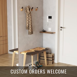 Könnte beinhalten: Eine Holzbank mit einer lebenden Kante und natürlicher Holzmaserung. Die Bank hat vier Beine und ist aus hellem braunem Holz gefertigt. Die Bank steht in einem Flur mit einer weißen Tür und einer weißen Wand. An einem Haken an der Wand hängt ein Schal. Der Text "CUSTOM ORDERS WELCOME" befindet sich auf dem Boden vor der Bank.
