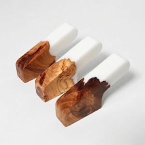 Op de afbeelding: Drie handgemaakte houten flessenstoppers. Elke stopper heeft een uniek houtnerfpatroon aan de basis, dat overgaat in een gladde, witte bovenkant. Het hout heeft warme bruine tinten en het witte gedeelte is een effen, ondoorzichtige kleur.