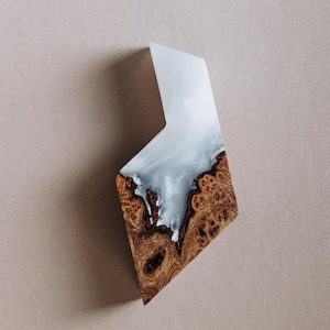Op de afbeelding: Een handgemaakte, geometrische hanger van hout en hars. Het onderste deel is van bruin wortelhout, het bovenste deel van helderwitte hars. Het midden heeft een wervelend, wolkachtig effect van wit en grijs. Ongeveer 10 cm lang.