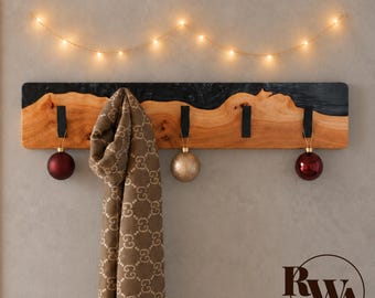 May include: A rectangular wall-mounted coat rack featuring a natural light wood grain edge combined with a dark black resin top. It includes four black metal hooks, each holding a round ornament, with a tan patterned scarf draped over one hook.