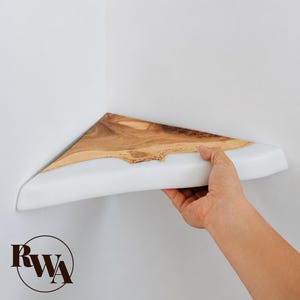 Puede incluir: Una estantería de esquina triangular que presenta una sección de grano de madera natural con un borde vivo y una sección de resina blanca opaca y lisa. La estantería está montada en una esquina y las letras RWA aparecen en un logo circular negro.