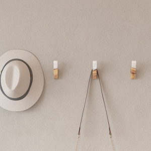 Op de afbeelding: Drie houten wandhaken met witte en houten accenten zijn op een neutraal gekleurde muur gemonteerd. Een strohoed met een donkergrijze band hangt links. Een zwarte handtas hangt aan de middelste haak.