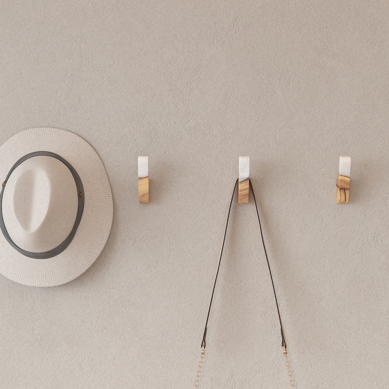 Op de afbeelding: Drie houten wandhaken met witte en houten accenten zijn op een neutraal gekleurde muur gemonteerd. Een strohoed met een donkergrijze band hangt links. Een zwarte handtas hangt aan de middelste haak.
