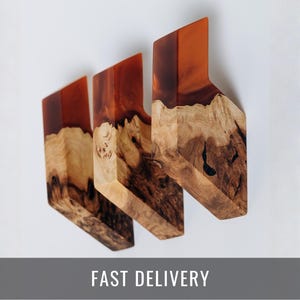May include: Three wooden cutting boards with a unique design. The top portion is a translucent orange resin, while the bottom is natural wood with dark brown and tan patterns. The boards are angled and have a modern aesthetic.